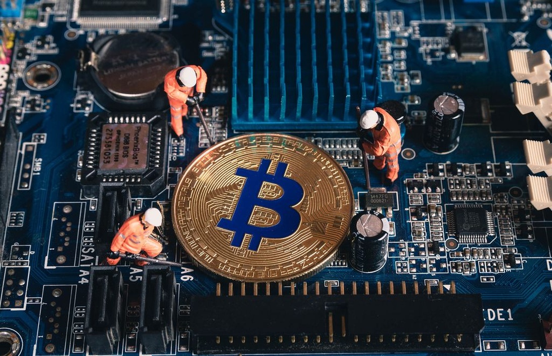 En kreativ miniatyrscen som visar små figurer av arbetare i orange säkerhetsutrustning som 'bryter' en fysisk Bitcoin placerad på ett datorkretskort.