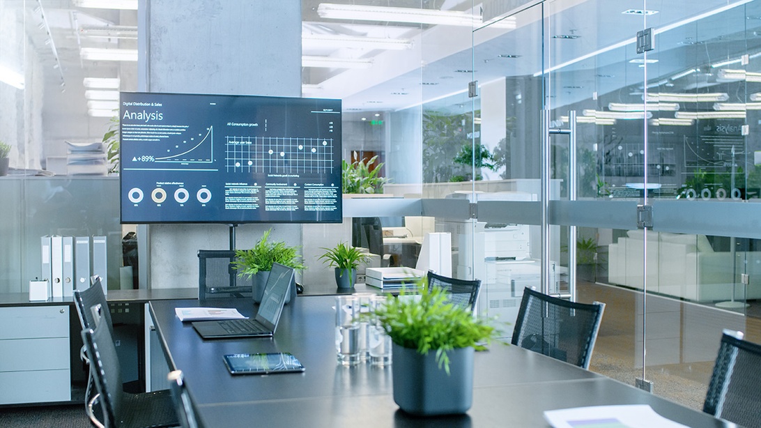 A sleek modern conference room featuring glass partitions, a dark conference table with laptops, and a large digital display showing business analytics dashboards with graphs and data visualization. The contemporary office space includes indoor plants for greenery and exemplifies the integration of technology in a modern workspace environment.