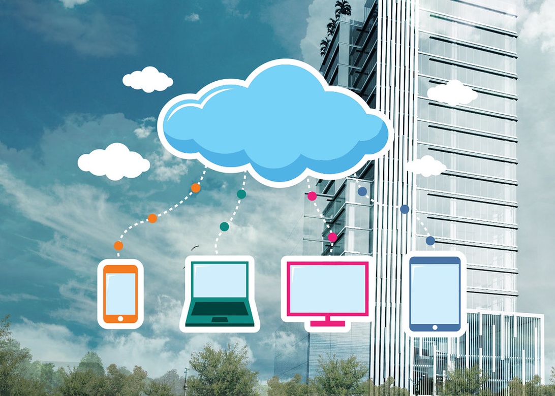 A conceptual illustration showing cloud computing technology with a blue cloud icon connected to various devices (smartphone, laptop, desktop monitor, and tablet). The background features a modern office building with glass architecture, representing how cloud technology enables connectivity in contemporary workplaces.