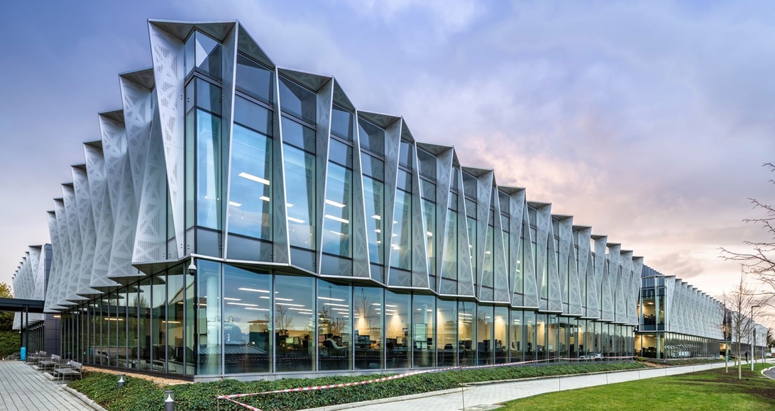 An exterior view of a contemporary office building with distinctive angular glass architecture. The structure features an innovative folded façade design with extensive glazing that allows natural light into the workspace. While representing a modern office environment, it doesn't directly showcase office technology equipment.